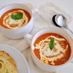 Bowl of roasted tomato vegetable soup topped with cream and fresh basil, served on a rustic wooden table.